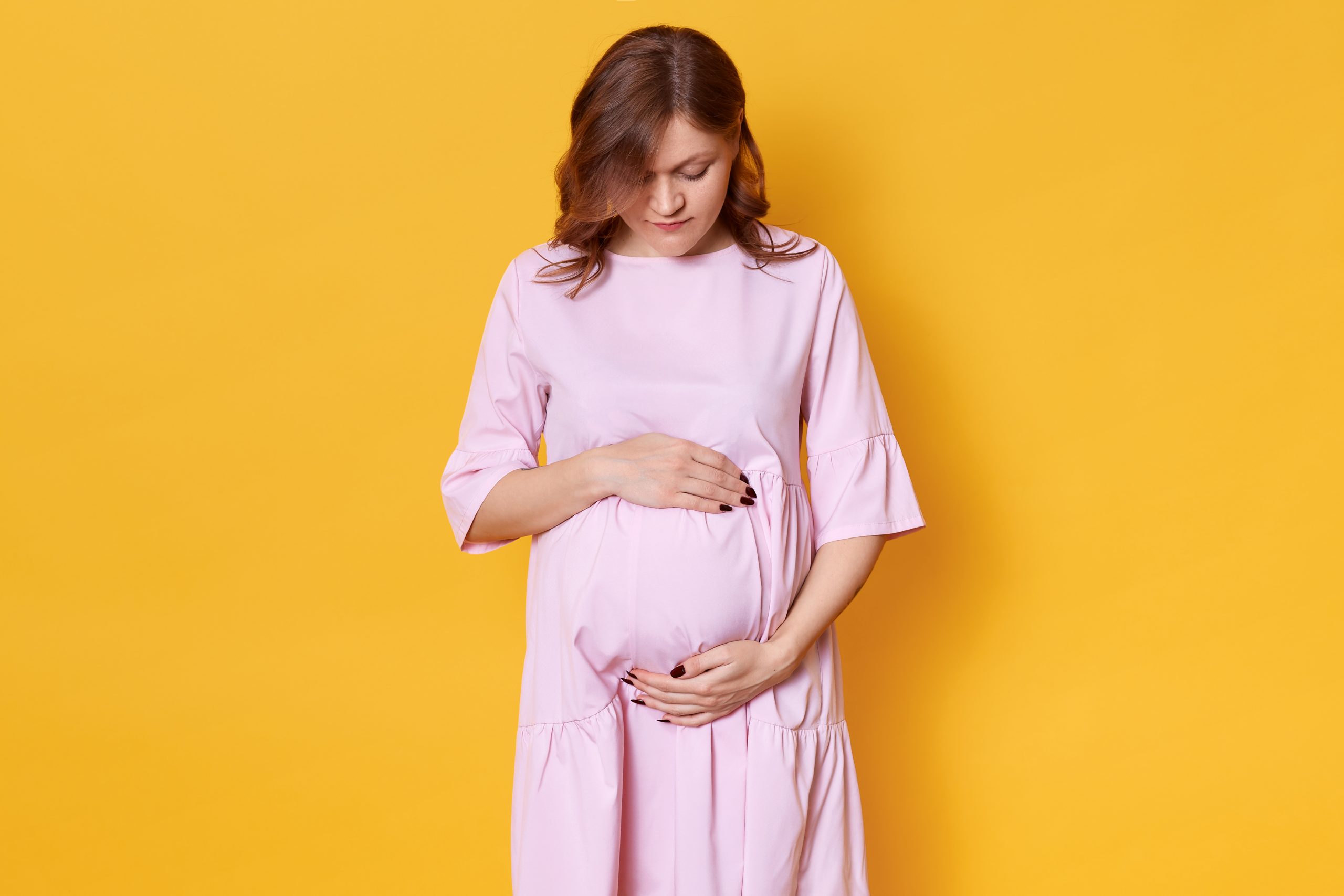 به طول کشیدن دوران زایمان Studio shot of young pregnant woman with brown hair, in elegant rose dress standing with hands on her belly in front of yellow background in studio, looking down on her abdomen, has dark manicure.