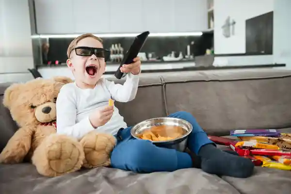 کودکی که عینک دارد و کنترل تلویزیون و یک کاسه چیپس